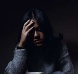 A woman sitting in darkness at a table, holding her head in discomfort with a mug on the table in front of her.