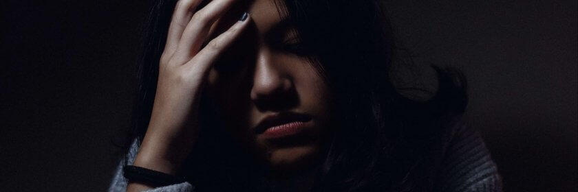 A woman sitting in darkness at a table, holding her head in discomfort with a mug on the table in front of her.