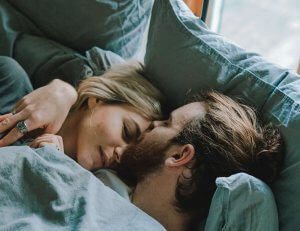 A young couple sleep in each others arms in a bed with blue pillows and blankets.