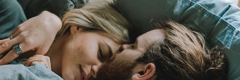 A young couple sleep in each others arms in a bed with blue pillows and blankets.