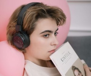 A young woman wearing headphones rests against a pink pillow while holding a book.