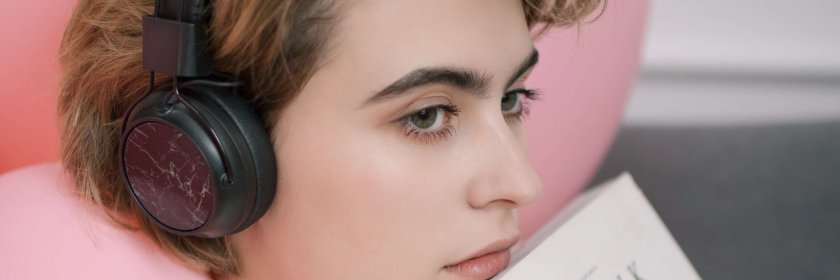 A young woman wearing headphones rests against a pink pillow while holding a book.